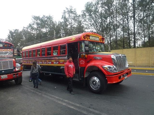 A red painted bus parked at the roadside, with two people walking beside it.